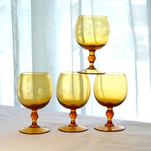 May include: Four amber-colored glass goblets with a round bowl and a stem. The goblets are arranged in a pyramid shape on a white surface.