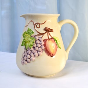 May include: A cream-colored ceramic pitcher with a handle, featuring a painted design of grapes and an apple.