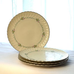 May include: A set of cream-colored porcelain plates with a floral design and gold trim. One plate stands upright, while three are stacked. The plates have a scalloped edge and a delicate pattern of pink and yellow flowers.