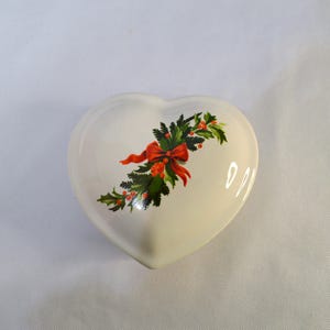 May include: A heart-shaped, off-white ceramic trinket box. The lid is decorated with a festive design of green holly leaves, red berries, and a red bow. The box is likely used for storing small jewelry items or keepsakes.
