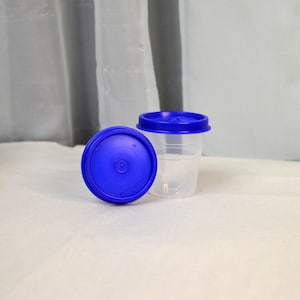 May include: Two clear plastic specimen cups with blue lids. One cup is upright with its lid on, and the other lid is off to the side. The cups are on a white surface.
