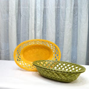 May include: Two oval-shaped baskets. One is a bright yellow, the other is olive green. Both have a lattice design and are made of plastic. The yellow basket is larger and has a slightly raised center.