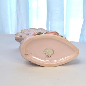 May include: A vintage, light pink ceramic vase with a scalloped edge and floral accents. The vase has a teardrop shape and a small, oval sticker on the bottom. The vase is decorated with pink and green flowers.