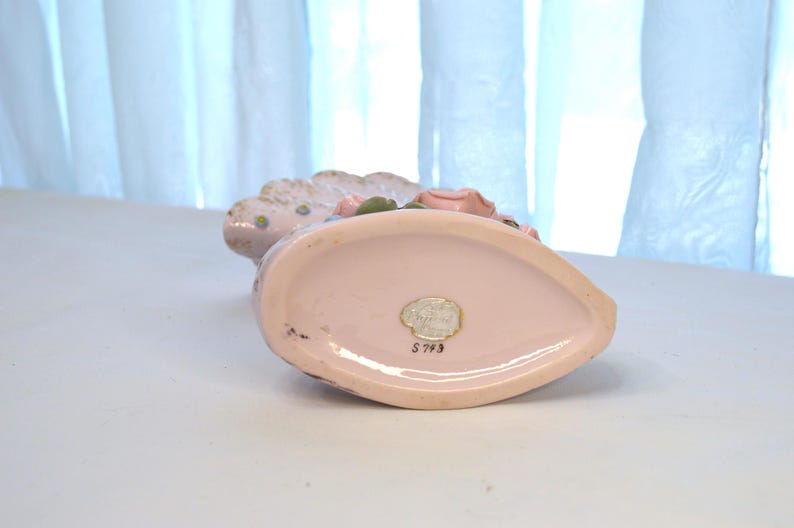 May include: A vintage, light pink ceramic vase with a scalloped edge and floral accents. The vase has a teardrop shape and a small, oval sticker on the bottom. The vase is decorated with pink and green flowers.