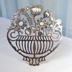 May include: Silver-plated decorative plate with a cut-out design of a vase with flowers. The vase is surrounded by a floral design with leaves and flowers.