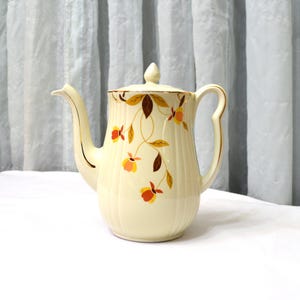 May include: Cream-colored vintage teapot with a lid and a curved handle. The teapot is decorated with a floral pattern in shades of brown, orange, and yellow. The spout and handle are accented with gold trim. The teapot is sitting on a white surface.