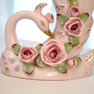 May include: A ceramic swan-shaped vase in soft pink, adorned with delicate pink roses and green leaves. The swan's beak is gold, and the vase has gold accents. A charming decorative piece.