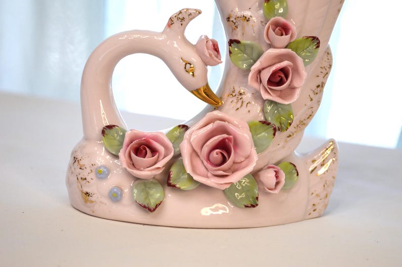 May include: A ceramic swan-shaped vase in soft pink, adorned with delicate pink roses and green leaves. The swan's beak is gold, and the vase has gold accents. A charming decorative piece.