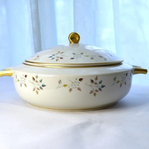 以下が含まれることがあります： 金色の縁取りと蓋が付いた白い磁器製のサービングディッシュ。ディッシュは、茶色、緑、青の葉の繊細な模様で飾られています。