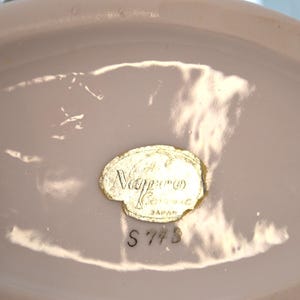 May include: A close-up of a pale pink ceramic object with a gold foil label. The label reads "Nippon Ceramic Japan" with the number "S 773" below. The object has a smooth, glossy finish.