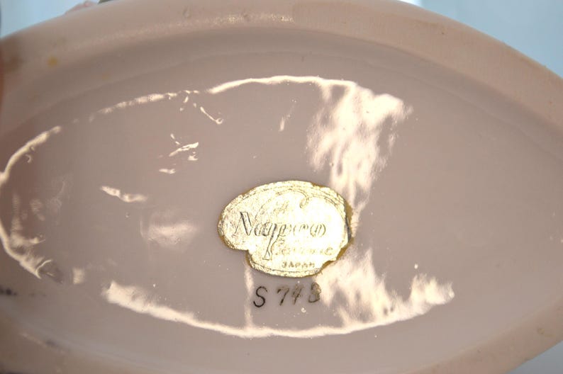 May include: A close-up of a pale pink ceramic object with a gold foil label. The label reads "Nippon Ceramic Japan" with the number "S 773" below. The object has a smooth, glossy finish.