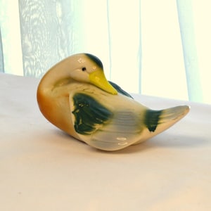 May include: A ceramic duck figurine with a white body, green and black wings, and a yellow beak. The duck is curled up in a sleeping position.