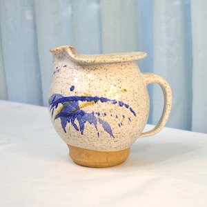 May include: A white ceramic pitcher with a speckled glaze and blue and yellow abstract design. The pitcher has a handle and a rounded body.