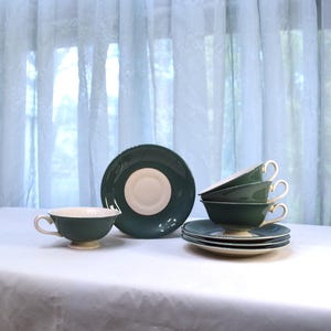 May include: A set of vintage teacups and saucers in a dark green color with cream-colored interiors and rims. The set includes teacups, saucers, and a larger plate, all arranged on a white surface. The teacups have delicate handles.