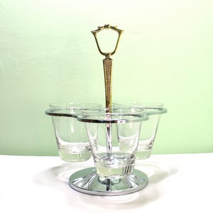 May include: A vintage chrome and gold-toned serving tray with four clear shot glasses. The tray has a handle and a circular base. The shot glasses have silver rims. The background is a light green color.