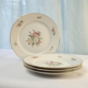 May include: Set of four white ceramic dinner plates with a floral design featuring pink and white roses. The plates have a gold rim.