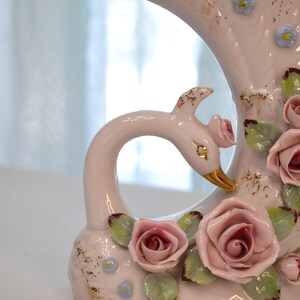 May include: A vintage ceramic swan vase in soft pink, adorned with delicate pink roses, green leaves, and gold accents. The swan's beak is gold. The vase has a curved design, perfect for floral arrangements.