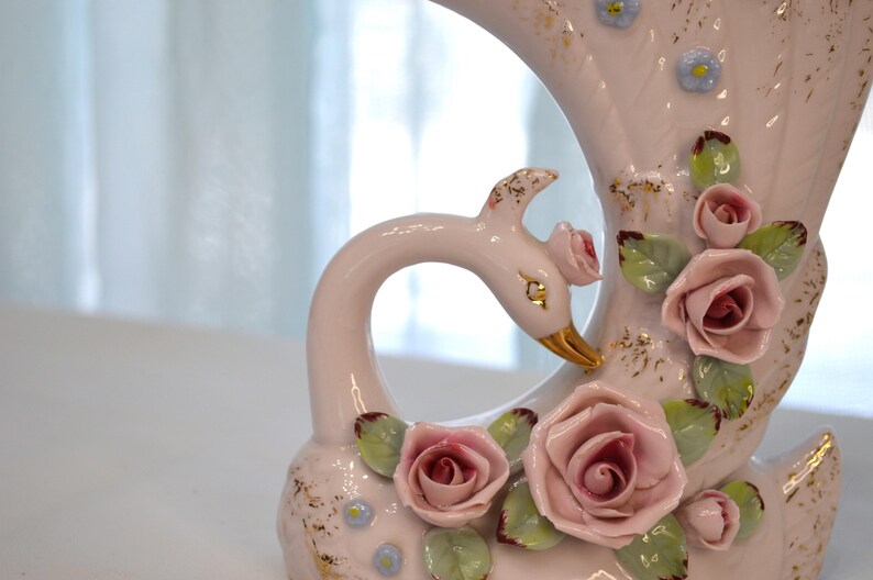 May include: A vintage ceramic swan vase in soft pink, adorned with delicate pink roses, green leaves, and gold accents. The swan's beak is gold. The vase has a curved design, perfect for floral arrangements.