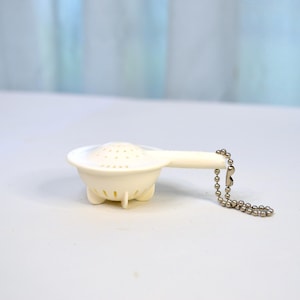 May include: A small, off-white tea infuser with a perforated bowl and a handle. It has three small feet and a chain with a ball-shaped design attached to the handle. The infuser is designed for steeping loose leaf tea.