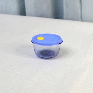 May include: A small, clear glass container with a blue lid and a yellow accent. The container is round and has a simple design. The lid fits snugly on top, creating an airtight seal. This container is ideal for storing small food items.