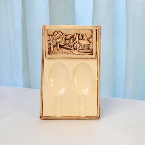 May include: A ceramic spoon rest with two slots for spoons. The rest is decorated with a raised image of a church and trees in a brown and white color scheme.
