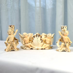 May include: A set of three vintage ceramic candle holders featuring cherub figures with gold accents. The center piece is a bowl with a leaf design.