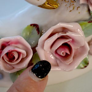 May include: Close-up of a white ceramic swan figurine adorned with pink and red roses and green leaves. The roses have detailed petals and are arranged around the swan's body. The leaves have red edges.