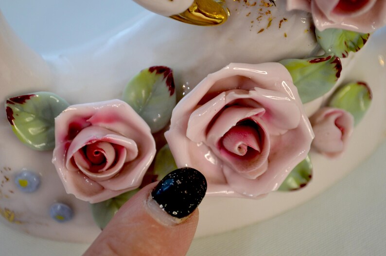 May include: Close-up of a white ceramic swan figurine adorned with pink and red roses and green leaves. The roses have detailed petals and are arranged around the swan's body. The leaves have red edges.