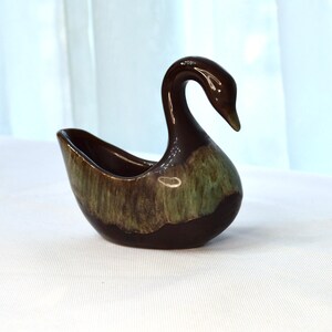 May include: A brown and green ceramic swan-shaped dish. The dish has a glazed finish and is perfect for holding small items.