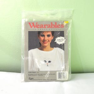 Puede incluir: Un kit de punto de cruz Wearables en un paquete de plástico transparente. La imagen muestra una camiseta blanca con un diseño de cara de gato. El texto "Wearables" es rojo, y el texto "Cat's Face" está debajo.