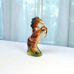 May include: A ceramic horse figurine in a rearing pose. The horse is primarily brown with a dark mane and tail, and a green base. The figurine has a glossy finish and is displayed on a white surface.