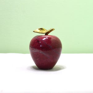 May include: A decorative apple figurine crafted from polished deep red stone with visible natural veining throughout the surface. The apple features a smooth, rounded body and is topped with a single, shiny metallic gold-toned leaf stem.