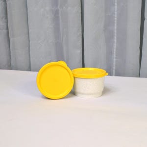 May include: A small, white container with a yellow lid, next to a separate yellow lid. The container is filled with small, white granules. The items are on a white surface, with a white curtain in the background.