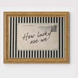 May include: A framed postcard with the text "How lucky are we." The postcard is set against a black and white striped background and framed in a light brown wooden frame. The postcard has a vintage aesthetic.