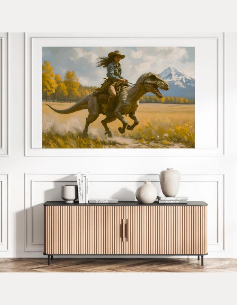 May include: A framed art print depicts a person riding a dinosaur through a field. The person wears a cowboy hat and denim shirt. The dinosaur is gray and brown. The background features a mountain, trees, and a yellow field.