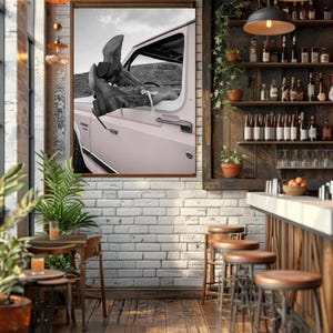 May include: A framed black and white photograph of cowboy boots resting on the open door of a pink vintage car. The image is displayed in a rustic bar setting with wooden stools, shelves of liquor bottles, and a brick wall.