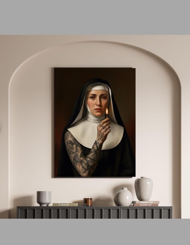 May include: A framed portrait of a nun in a black habit and white wimple, holding a lit match. The nun has a full sleeve tattoo on her arm. The background is a dark red. The portrait is hanging on a beige wall.