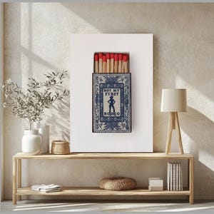 May include: A framed print featuring a blue matchbox design with the text "NOT MY FIRST RODEO". The matchbox has a decorative border and a silhouette. The matches inside have red tips.