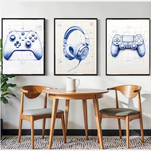 May include: Three framed blue and white gaming-themed art prints. The prints feature detailed illustrations of a game controller, headphones, and another game controller, all rendered in a blueprint style. The artwork is displayed above a wooden table and chairs.