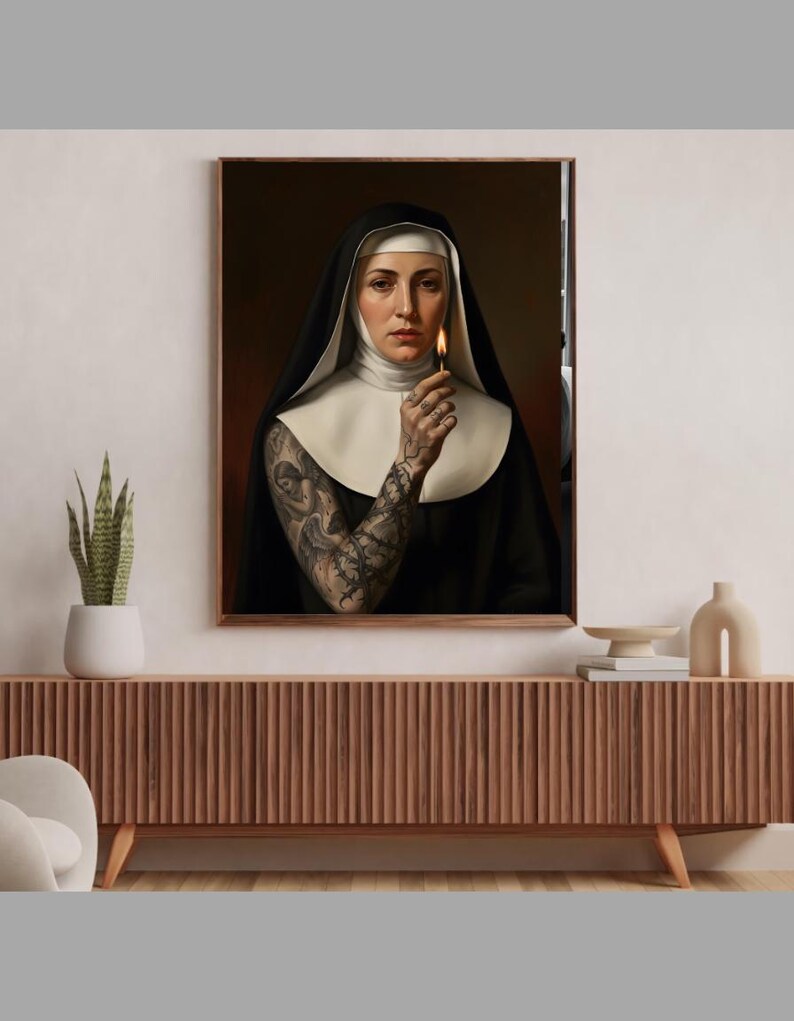 May include: A framed portrait of a nun holding a lit match. The nun is wearing a traditional black habit with a white wimple and collar. Her arm is covered in intricate tattoos. The artwork is displayed on a wooden cabinet.
