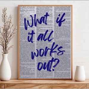 Puede incluir: Una impresión enmarcada con el texto "What if it all works out?" en escritura azul oscuro. El fondo es una página de libro vintage. El marco es de color marrón claro. La impresión se muestra sobre una superficie de madera.