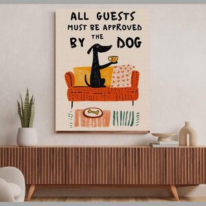 以下が含まれることがあります： オレンジ色のソファに座り、カップを持った黒い犬が描かれた、気まぐれなウォールアートプリントです。「ALL GUESTS MUST BE APPROVED BY THE DOG」と書かれています。アートワークには骨の形をしたお皿とカラフルなアクセントが含まれています。