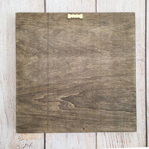 Puede incluir: Letrero cuadrado de madera con tinte marr&oacute;n oscuro, con veta de madera visible. Un peque&ntilde;o colgador de dientes de sierra dorado est&aacute; fijado en la parte superior central para montaje en pared. El letrero se muestra sobre un fondo de madera clara.