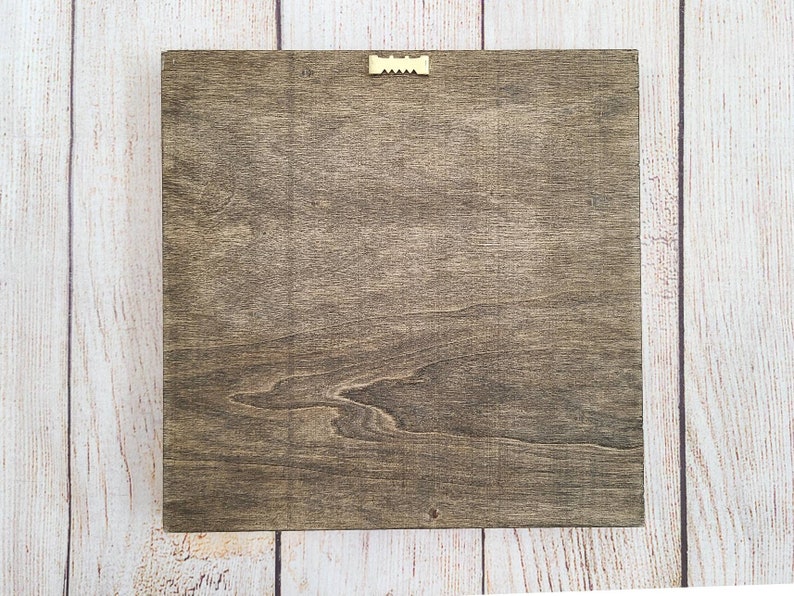 Puede incluir: Letrero cuadrado de madera con tinte marr&oacute;n oscuro, con veta de madera visible. Un peque&ntilde;o colgador de dientes de sierra dorado est&aacute; fijado en la parte superior central para montaje en pared. El letrero se muestra sobre un fondo de madera clara.