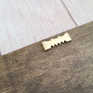 Puede incluir: Primer plano de una secci&oacute;n de un marco de madera con un colgador de dientes de sierra dorado. La madera tiene un aspecto natural y envejecido con vetas visibles y un color marr&oacute;n apagado. El colgador est&aacute; bien sujeto a la madera.