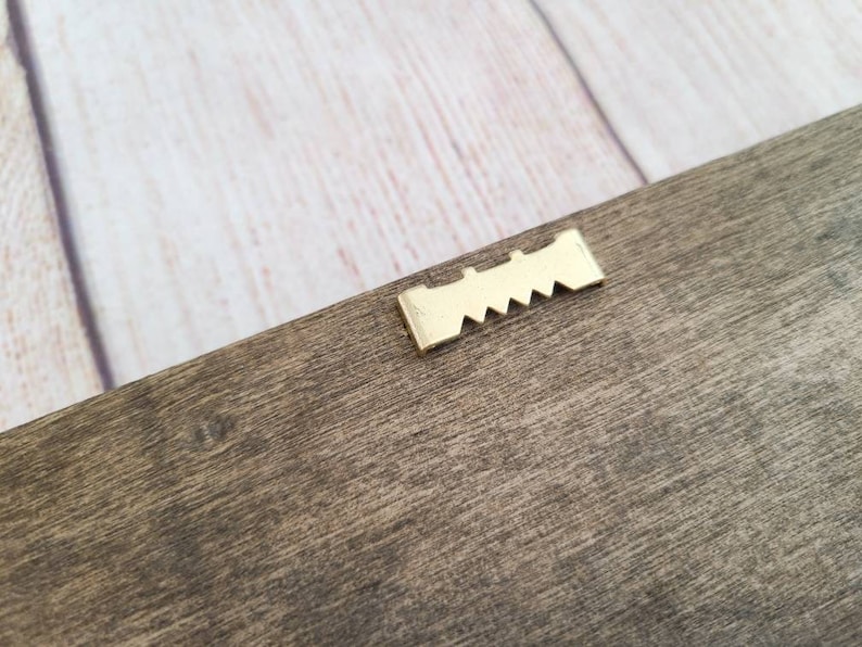 Puede incluir: Primer plano de una secci&oacute;n de un marco de madera con un colgador de dientes de sierra dorado. La madera tiene un aspecto natural y envejecido con vetas visibles y un color marr&oacute;n apagado. El colgador est&aacute; bien sujeto a la madera.