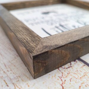 Puede incluir: Un letrero de madera enmarcado con un acabado marr&oacute;n oscuro desgastado. El letrero presenta un fondo blanco con texto negro, parcialmente visible. El marco es cuadrado y la veta de la madera es visible.