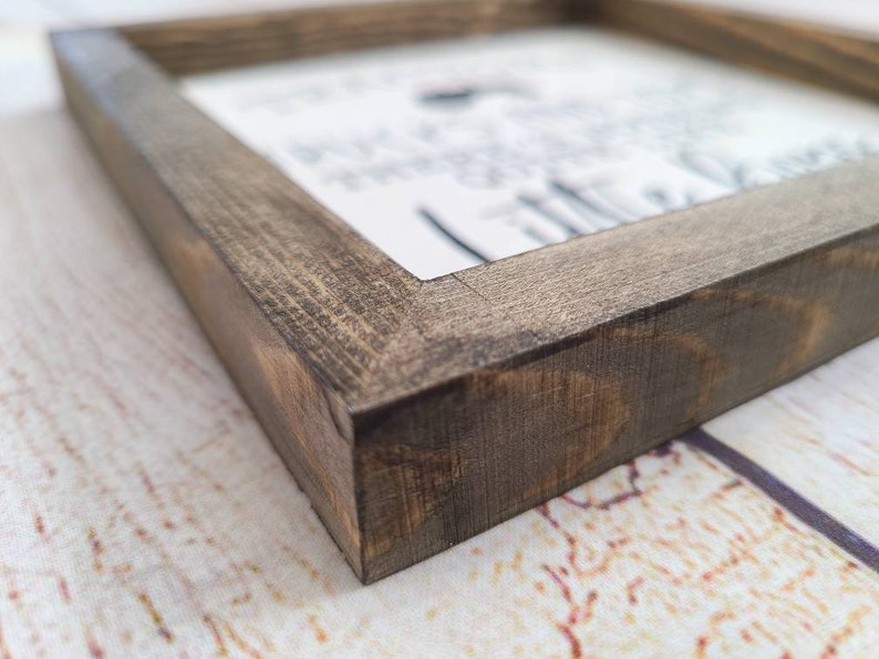 Puede incluir: Un letrero de madera enmarcado con un acabado marr&oacute;n oscuro desgastado. El letrero presenta un fondo blanco con texto negro, parcialmente visible. El marco es cuadrado y la veta de la madera es visible.