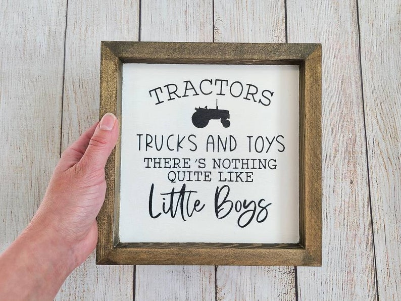Puede incluir: Letrero cuadrado con marco de madera con el texto "Tractors, Trucks and Toys, There's Nothing Quite Like Little Boys" en letras negras. El letrero tiene un fondo blanco y un marco marr&oacute;n oscuro.