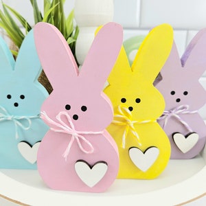 May include: Four wooden bunny figurines in pastel colors, blue, pink, yellow, and purple. Each bunny has a white heart attached to the bottom and a white ribbon tied around the middle.
